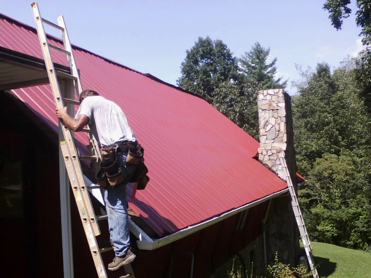 Roofer on a ladder beside a red metal roof and stone chimney — Soffit & Fascia Repair by Roofing Brothers in Waban